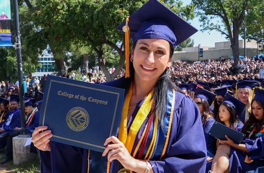 A female student in cap and gown smiles while holding up her degree during commencement ceremony
